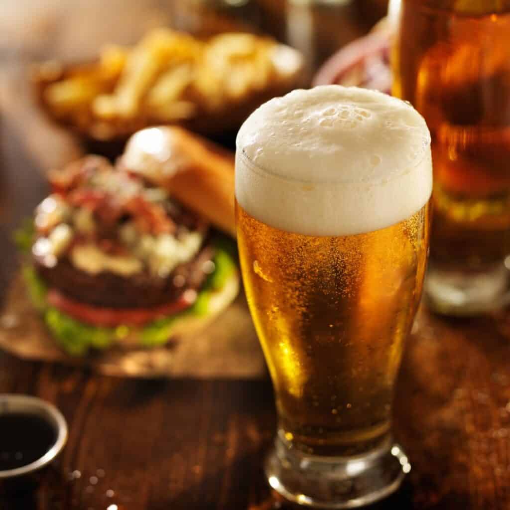 a glass of beer and a hamburger which is blurry on a wooden table