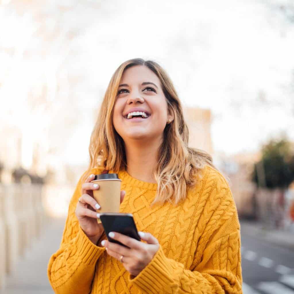 a person in a yellow sweater is smiling while holding a cell phone and coffee