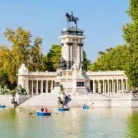 A photo of rowboats on the Buen Retiro Park lake in Madrid, Spain, with the equestrian Monument to Alfonso XII in the background.