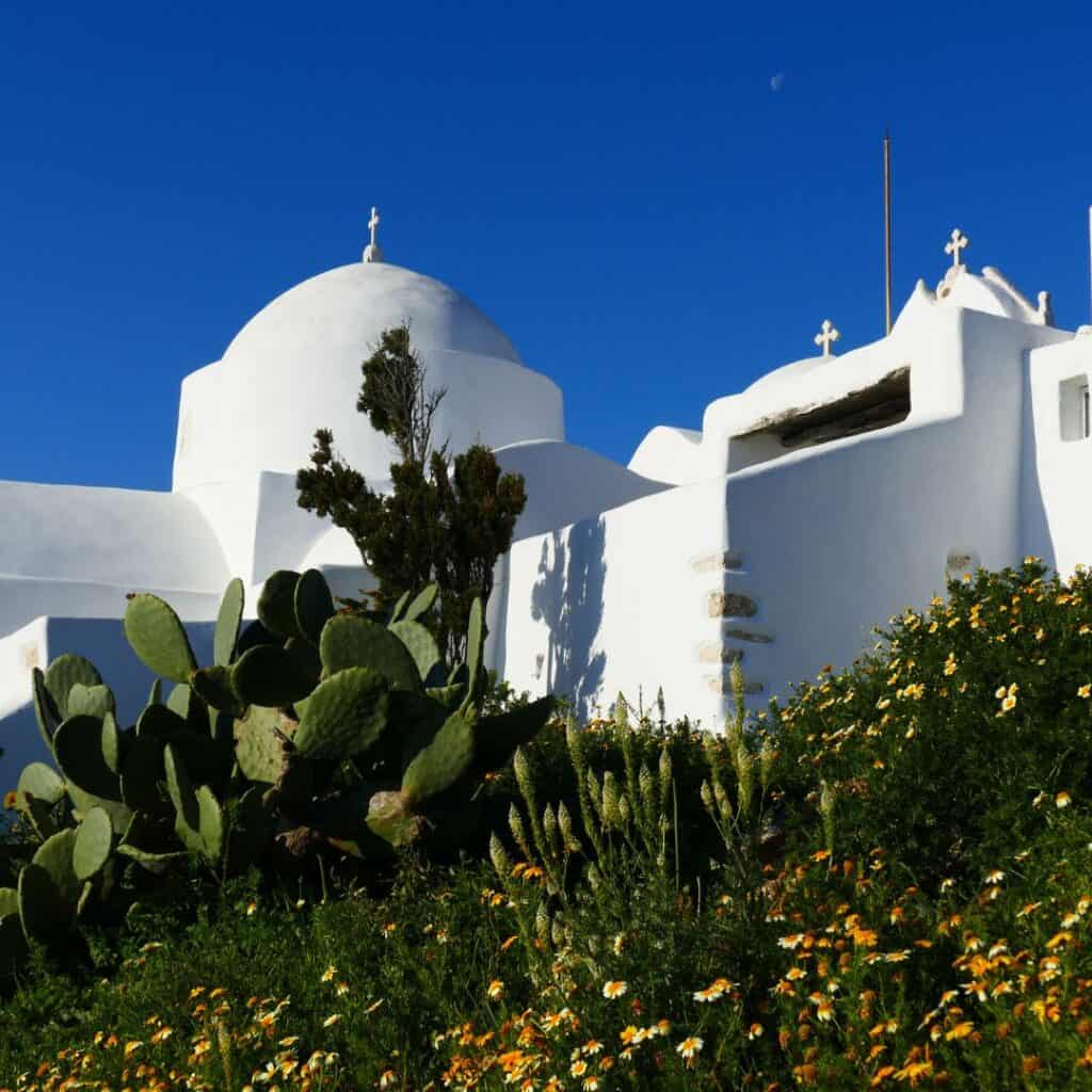 A picturesque scene of Agios Antonios Monastery on the Greek island of Paros, featuring whitewashed buildings, a lush green cactus, vibrant yellow flowers, and a crescent moon in the daytime sky.