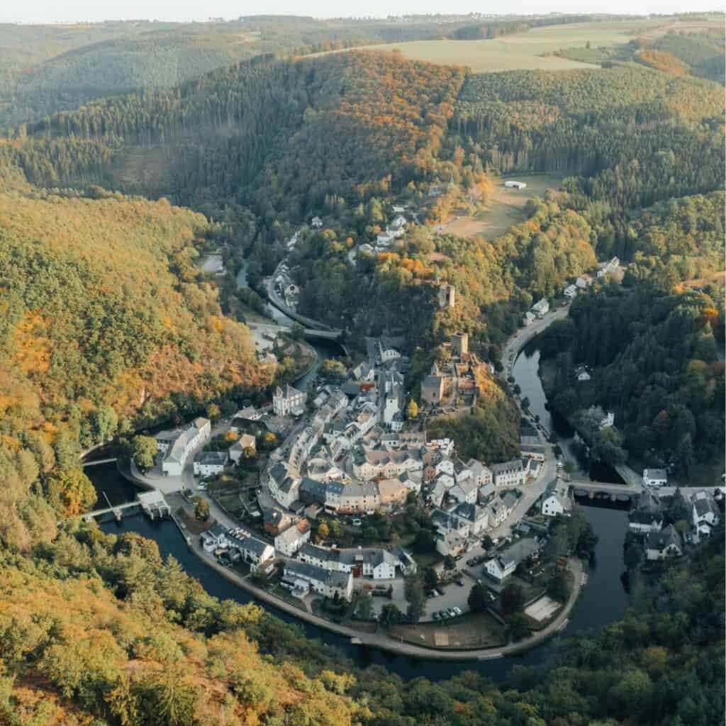 An aerial view of Esch-sur-Sûre, nestled among trees with a river winding through it. This town is located along the Autopedestre Esch-sur-Sure Trail in Luxembourg.