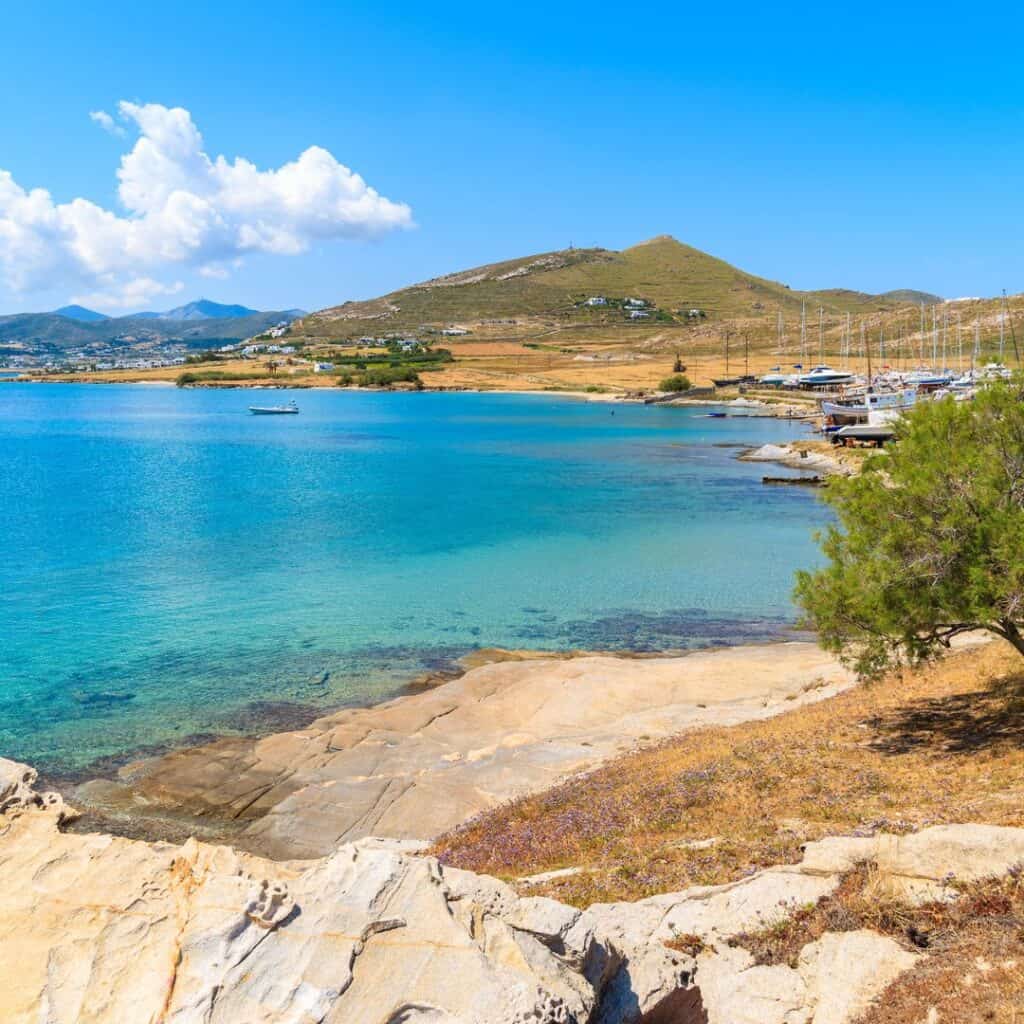 A stunning panoramic view of Monastiri Beach in Paros, featuring a vibrant blue sky, fluffy white clouds, a lush green tree, and a rocky shoreline leading to calm, turquoise waters with sailboats anchored in the bay.