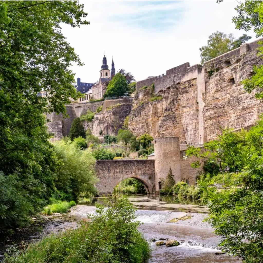 A stone bridge with multiple arches crosses a river in front of a large, ancient castle with round towers. The castle is part of the Bock Casemates, a network of fortified tunnels that are now a popular tourist destination in Luxembourg City.