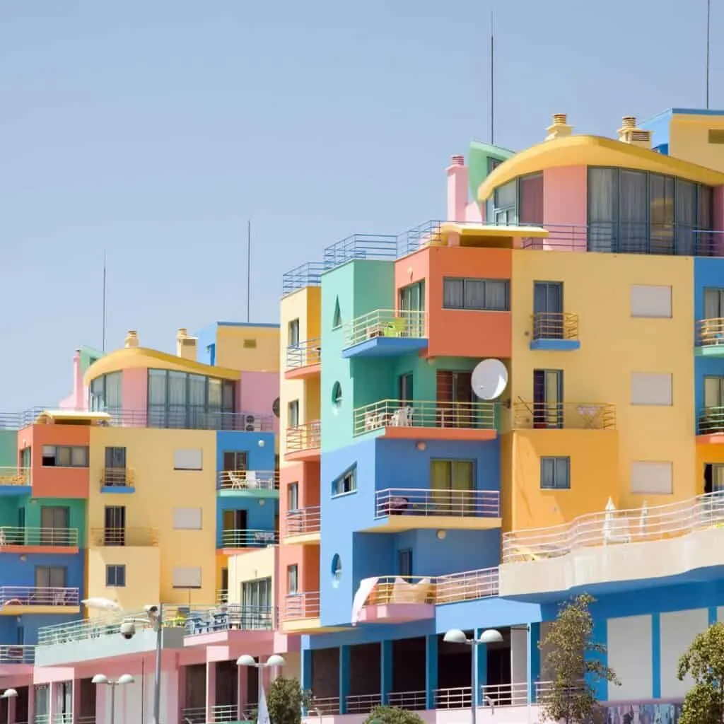 A row of brightly colored buildings with balconies overlooking the Albufeira Marina in Portugal. There are boats moored in the marina and palm trees lining the waterfront. The sky is clear and blue.