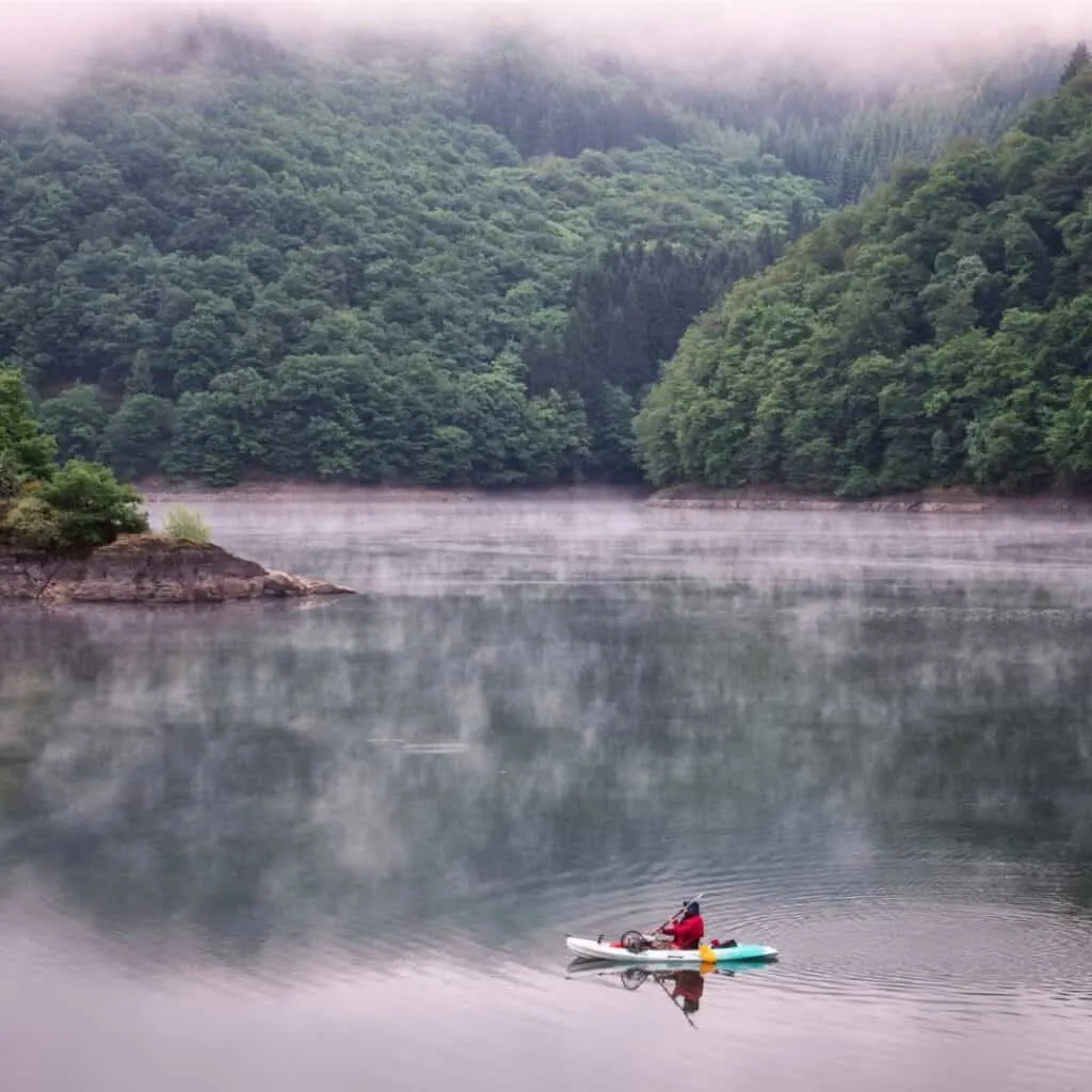 A serene scene of a lone kayaker gliding across the tranquil waters of Lac de la Haute-Sûre, with the verdant hills of Luxembourg's Ardennes region providing a picturesque backdrop.