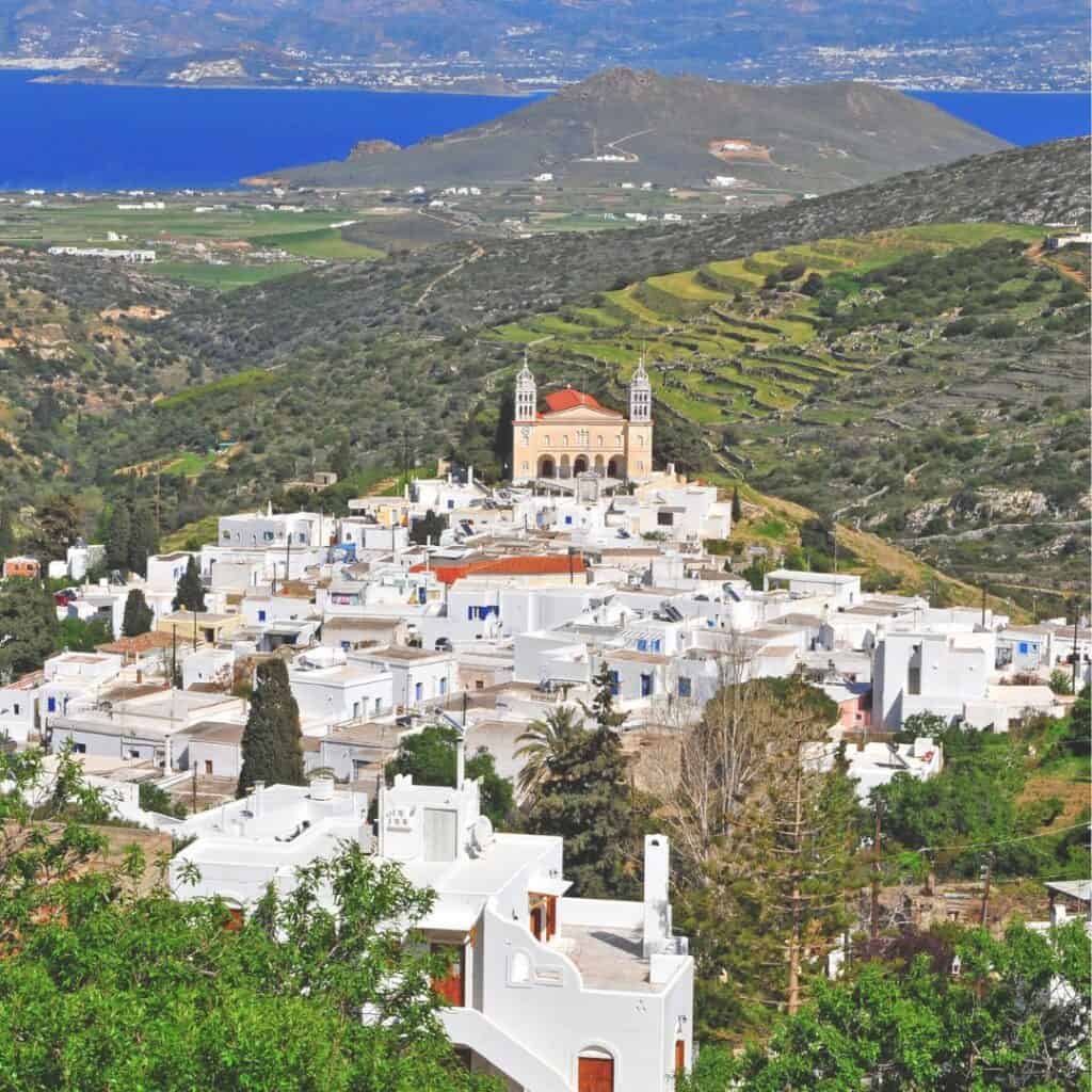 A breathtaking aerial view of Lefkes Village on the Greek island of Paros, showcasing a traditional Cycladic settlement with whitewashed houses, a prominent church with twin bell towers, and a panoramic vista of the Aegean Sea.