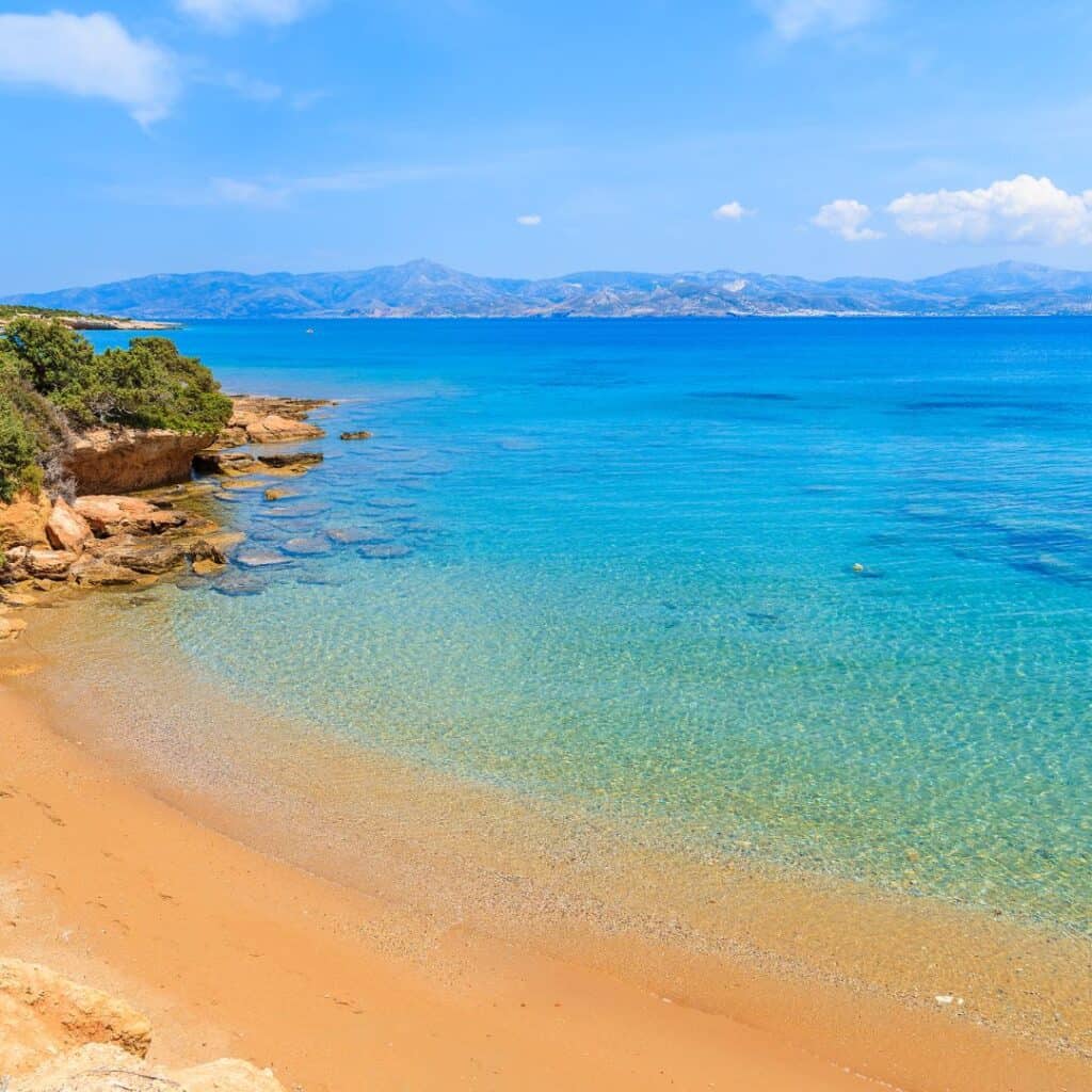 A stunning panoramic view of Santa Maria Beach in Paros, featuring a long stretch of golden sand, gentle waves lapping at the shore, and a small red boat bobbing in the calm waters, with a backdrop of whitewashed buildings and lush green hills under a clear blue sky.