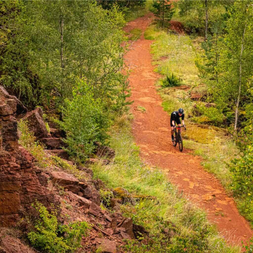 A person on a bicycle rides down a scenic dirt road amidst a lush green forest. The Minett Trail, a 90-kilometer hiking and biking path in southern Luxembourg, winds through the heart of the region's former industrial landscape, offering stunning views of rolling hills, forests, and rock formations.