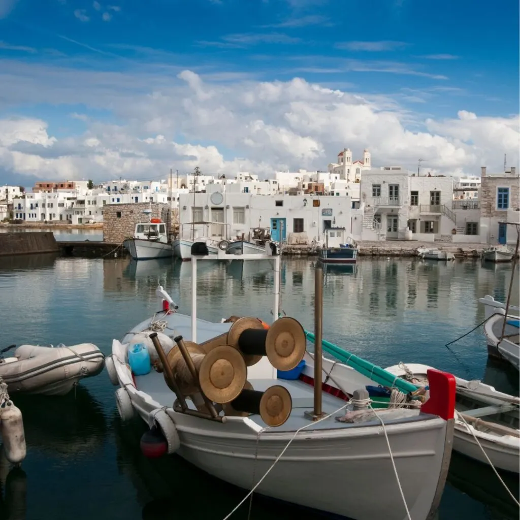 Naoussa Harbor with fishing boats on the island of Paros, Greece. In the background are white Cycladic architecture underneath a blue sky with white clouds.