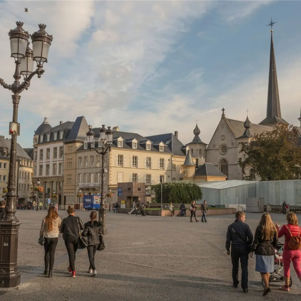 Photo of Place Guillaume II, featuring a equestrian statue of Grand Duke Guillaume II in the center. The surrounding area is filled with trees and people shopping, which is one of the fun things to do in Luxembourg City. In the background, the towers of the Cathédrale Notre-Dame can be seen.