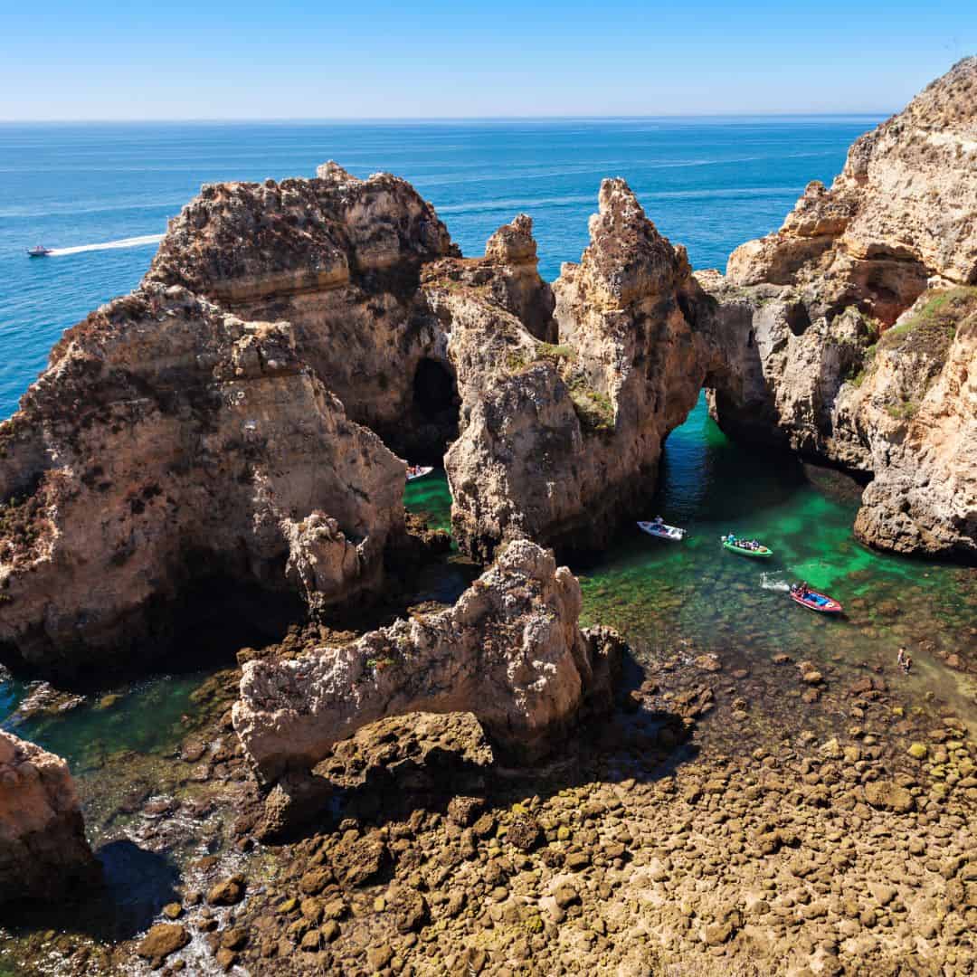 An aerial view of Ponta da Piedade, a rocky coastline in Portugal with turquoise water and several boats. The rocks are formed by millions of years of erosion and have a variety of shapes and sizes. The water is crystal clear and you can see the bottom of the ocean floor. The sky is blue and there are a few white clouds.