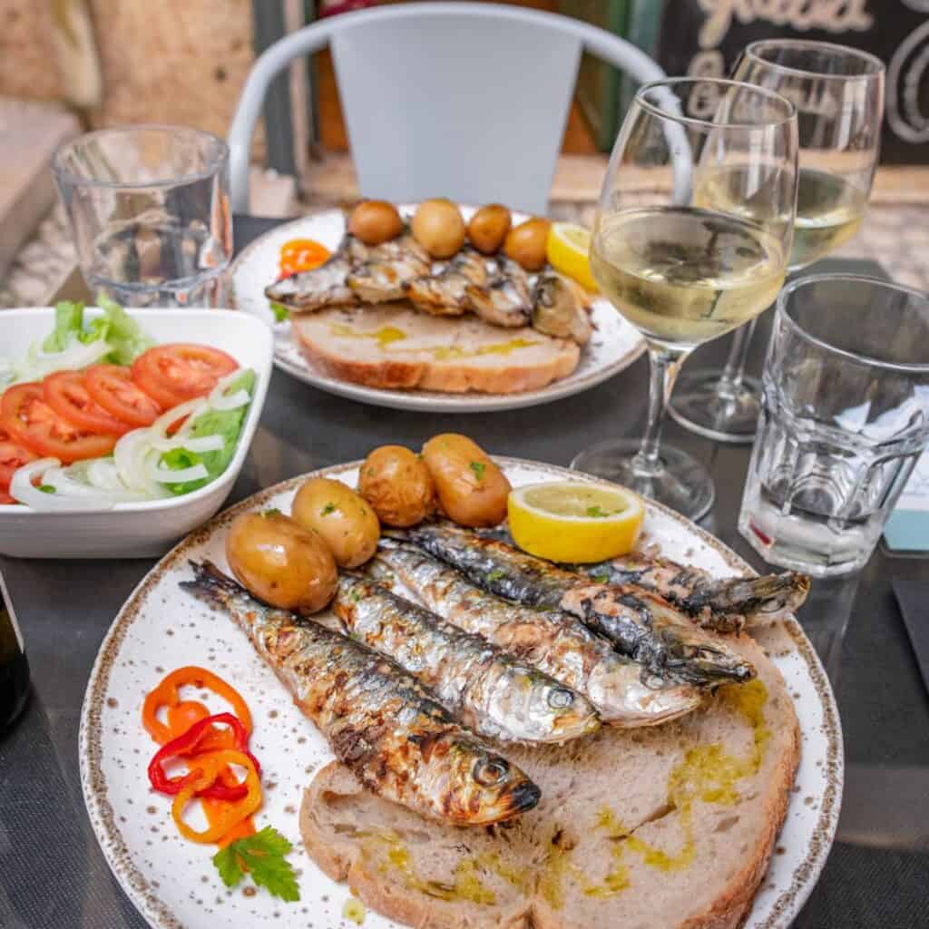 A plate of grilled sardines, potatoes, and salad with a glass of white wine served in a restaurant in Olhao. The sardines are served on a bed of bread and garnished with lemon wedges and red pepper slices. The photo is taken from a close-up perspective, with the sardines in focus.