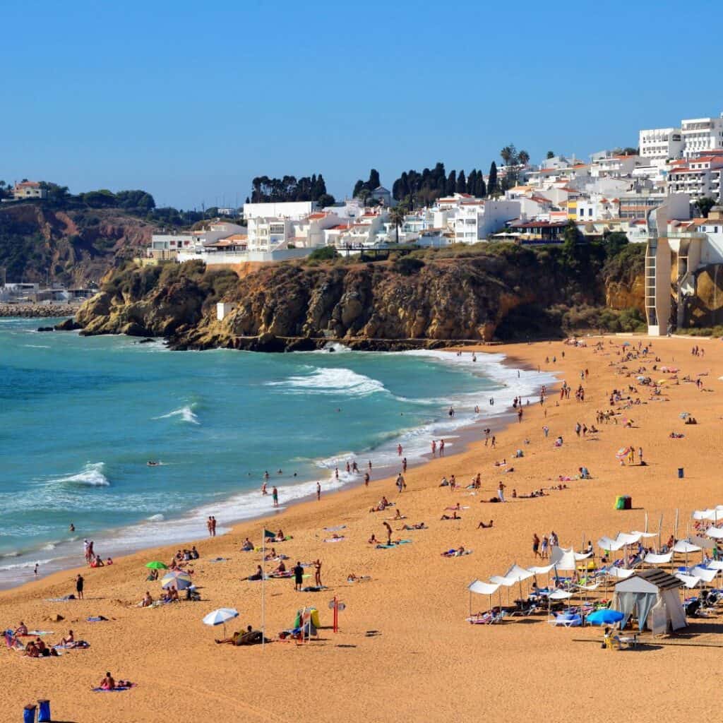 A photo of Praia dos Pescadores beach in Albufeira, Portugal. The beach is lined with colorful umbrellas, lounge chairs, and people sunbathing. In the background, there are cliffs and buildings.