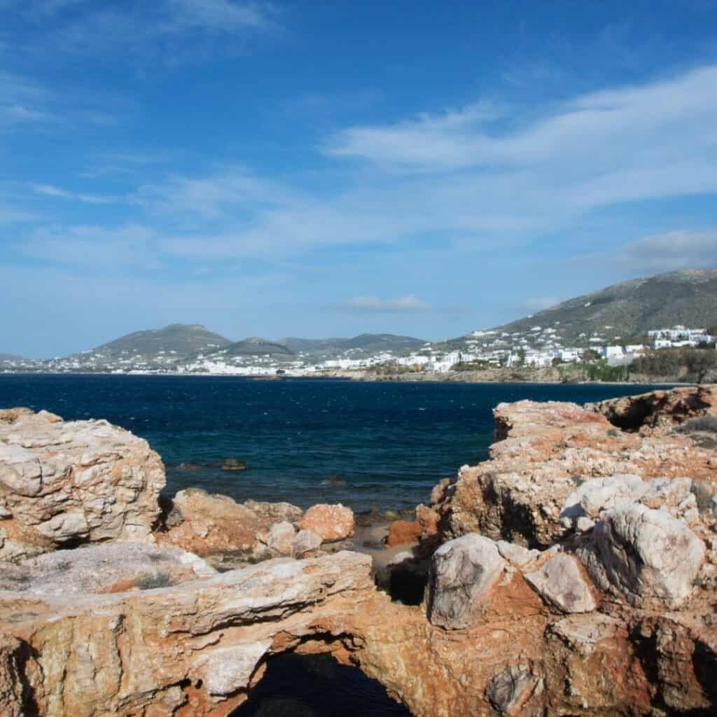 A scenic view of Paros Park, featuring a rocky coastline with a natural arch formation, overlooking the picturesque town of Paros with its whitewashed buildings and clear blue sea.