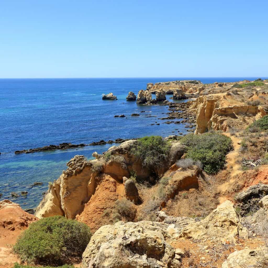 Scenic view of the Algarve coastline from the Caminho da Baleeira Nature Reserve. The image shows rugged cliffs overlooking Praia da Arrifes, a small beach with blue water and white sand.