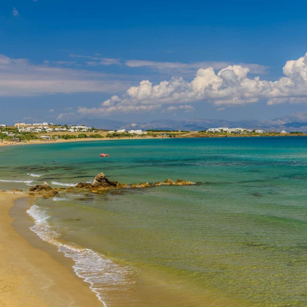 A picturesque scene of Golden Beach in Paros, featuring a long stretch of golden sand, gentle waves lapping at the shore, and a small red boat bobbing in the calm waters, with a backdrop of whitewashed buildings and lush green hills under a clear blue sky.