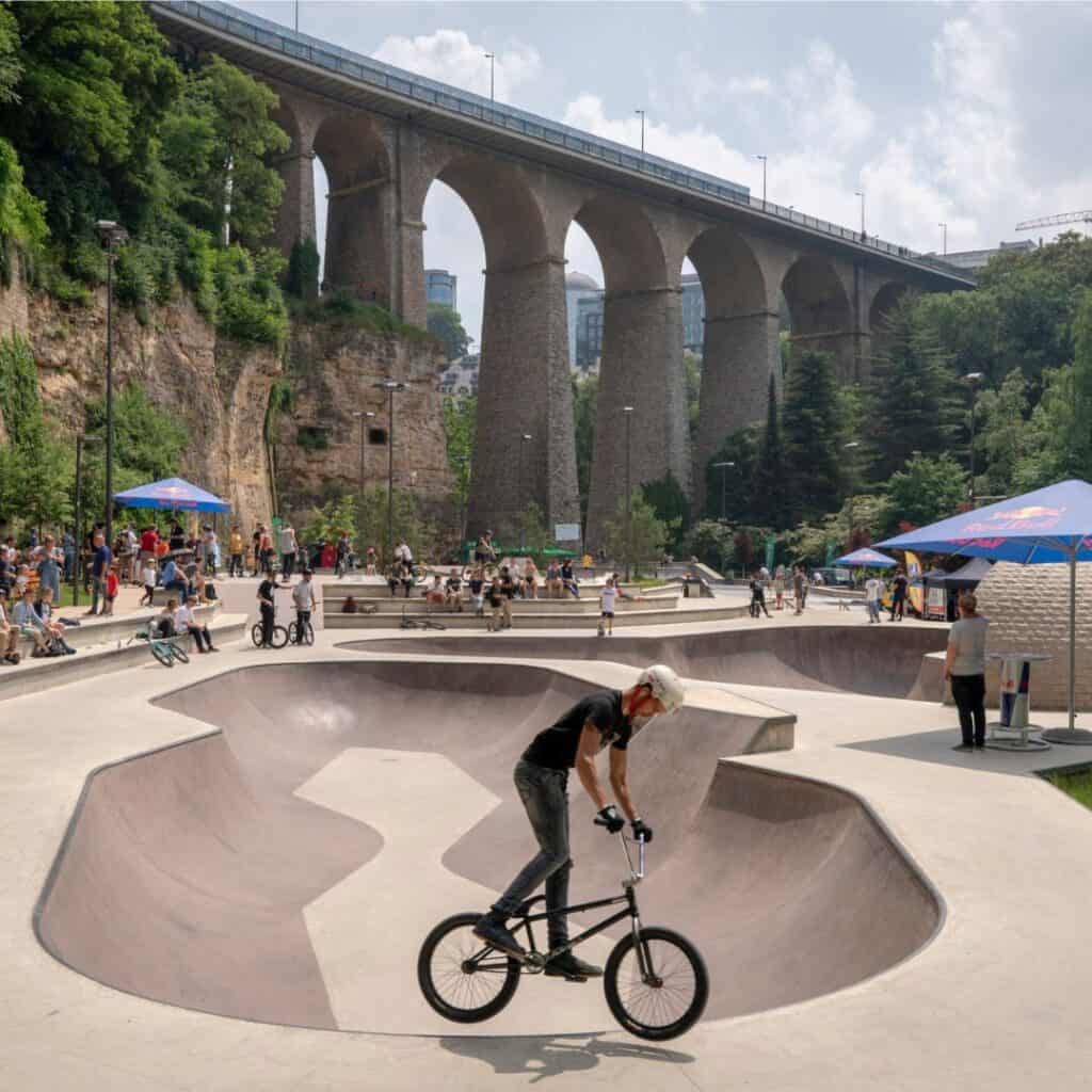 A vibrant aerial photograph capturing the sprawling layout of Skatepark Péitruss, set against the lush greenery of the Pétrusse Valley in Luxembourg City. The skate park's diverse array of ramps, rails, and bowls are clearly visible, showcasing its versatility for various skating disciplines. In the background, the historic fortifications of the city's old town stand as a testament to Luxembourg's rich heritage.