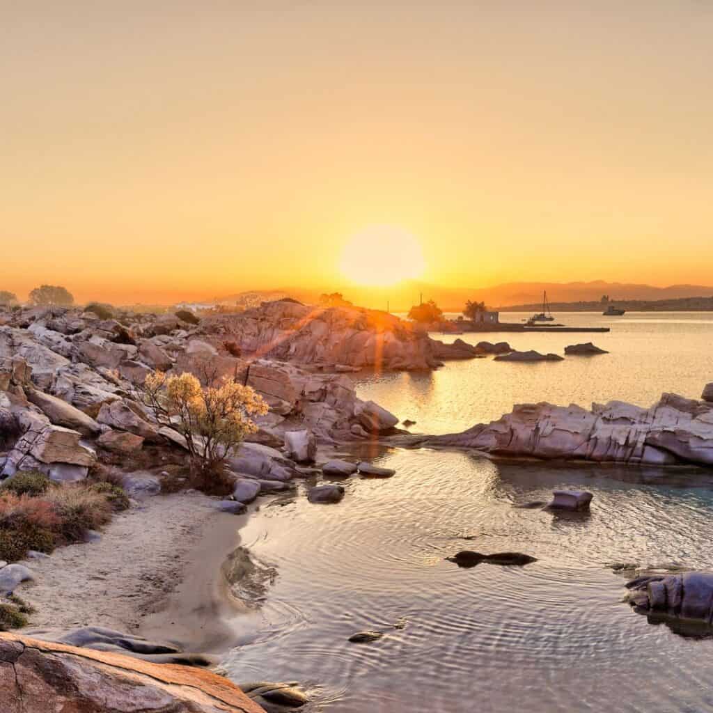 A stunning sunrise illuminates the rocky coastline of Kolymbithres Beach on the Greek island of Paros. The calm water reflects the warm colors of the sky.