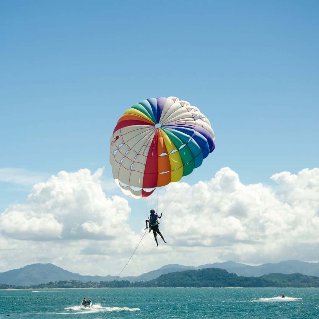 A person parasailing over a blue ocean in Albufeira, Portugal. The person is in a brightly colored parachute harness attached to a long tow rope behind a boat.