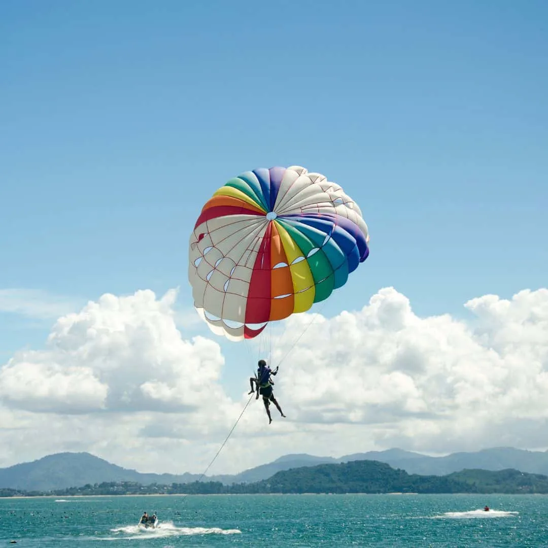 A person parasailing over a blue ocean in Albufeira, Portugal. The person is in a brightly colored parachute harness attached to a long tow rope behind a boat.