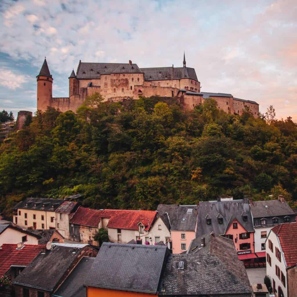 Vianden Castle, a large stone castle with round towers, sits on a hilltop overlooking a small town in Luxembourg. The castle is surrounded by trees.