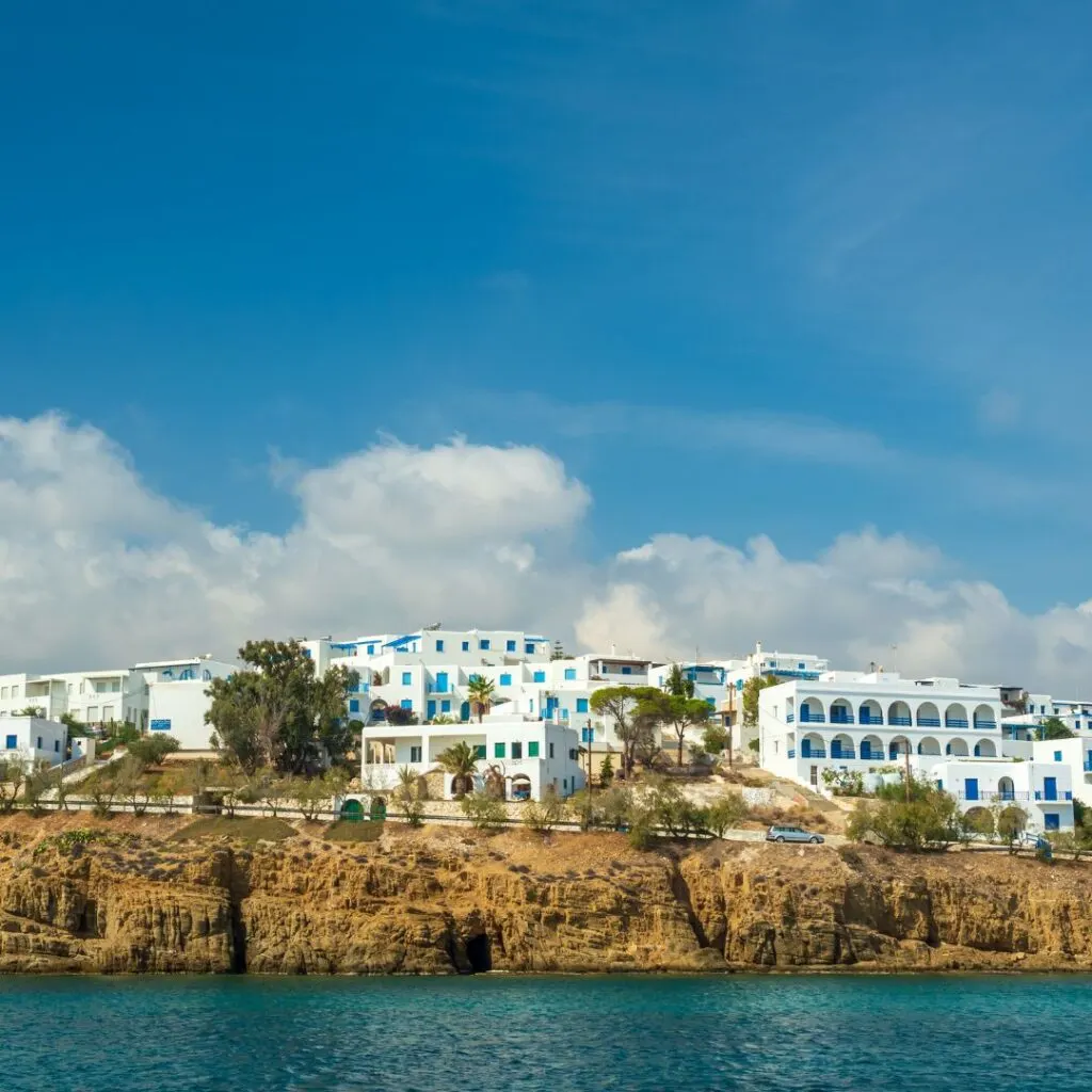 A picturesque view of Piso Livadi, Paros, showcasing whitewashed buildings perched on a cliffside overlooking the crystal-clear blue sea under a vibrant sky with fluffy clouds.