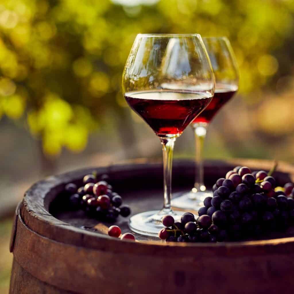 Two glasses of red wine and a bunch of grapes on a wooden barrel in a vineyard in Algarve. The sun is setting in the background, and the sky is a deep blue. The image is taken from a close-up perspective, and the focus is on the wine and the grapes.