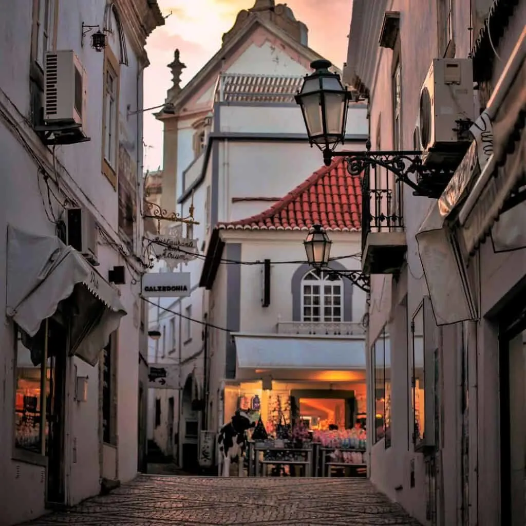 A photo of a narrow cobblestone street lined with whitewashed buildings with red tile roofs in the old town of Albufeira, Portugal. There are a few signs in Portuguese visible on the buildings, including one for a restaurant and a clothing store. There are also a few people walking down the street.