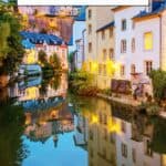 A Pinterest pin about Luxembourg Bucketlist showing a scenic overlook of Luxembourg City at sunset, with colorful buildings nestled along the valley sides.