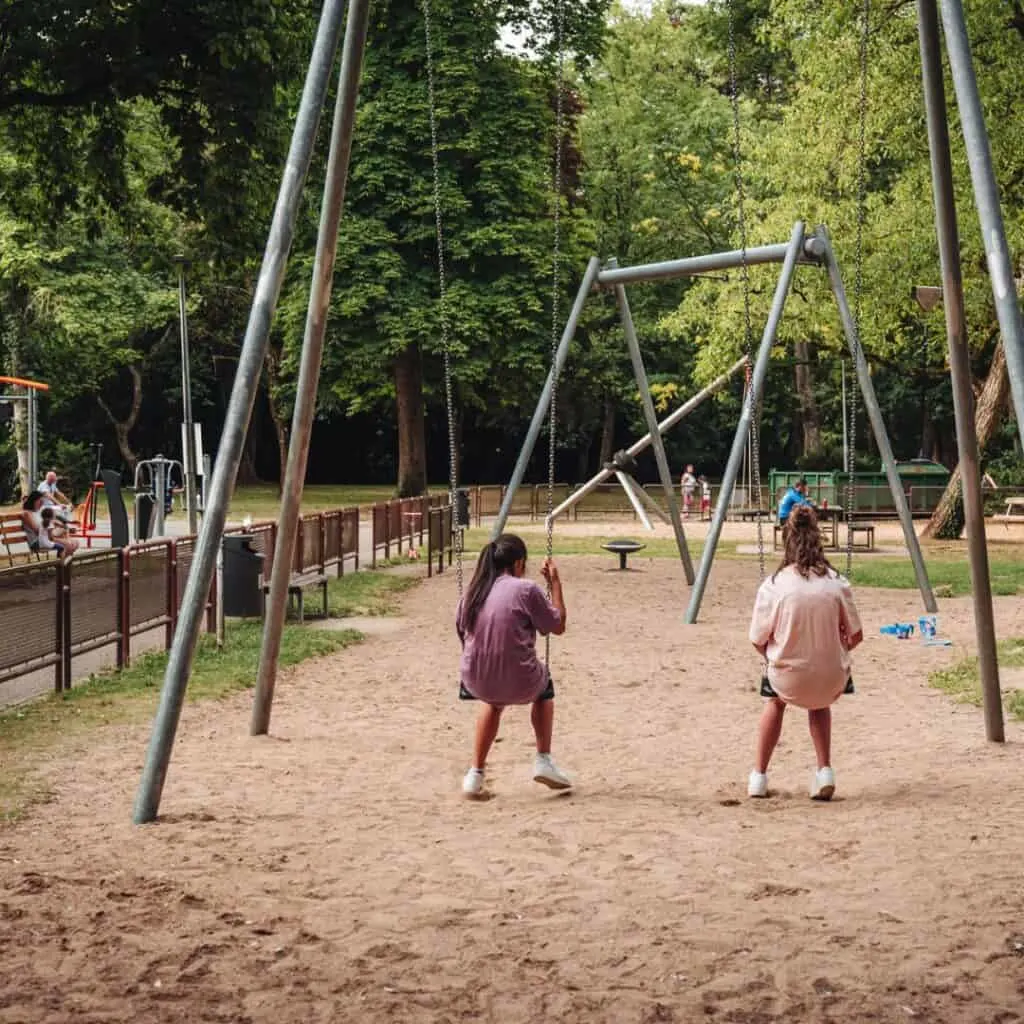 Two girls sitting on swings in a sandy playground at Parc Merveilleux in Luxembourg, surrounded by trees and playground equipment.