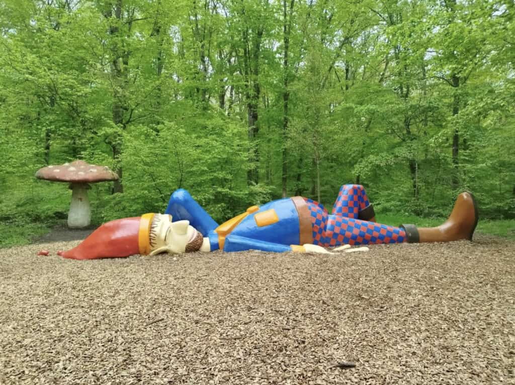 Colorful giant gnome sculpture lying down on wood chips in a lush green forest at Merveilleux Park, Luxembourg, with a large mushroom structure in the background