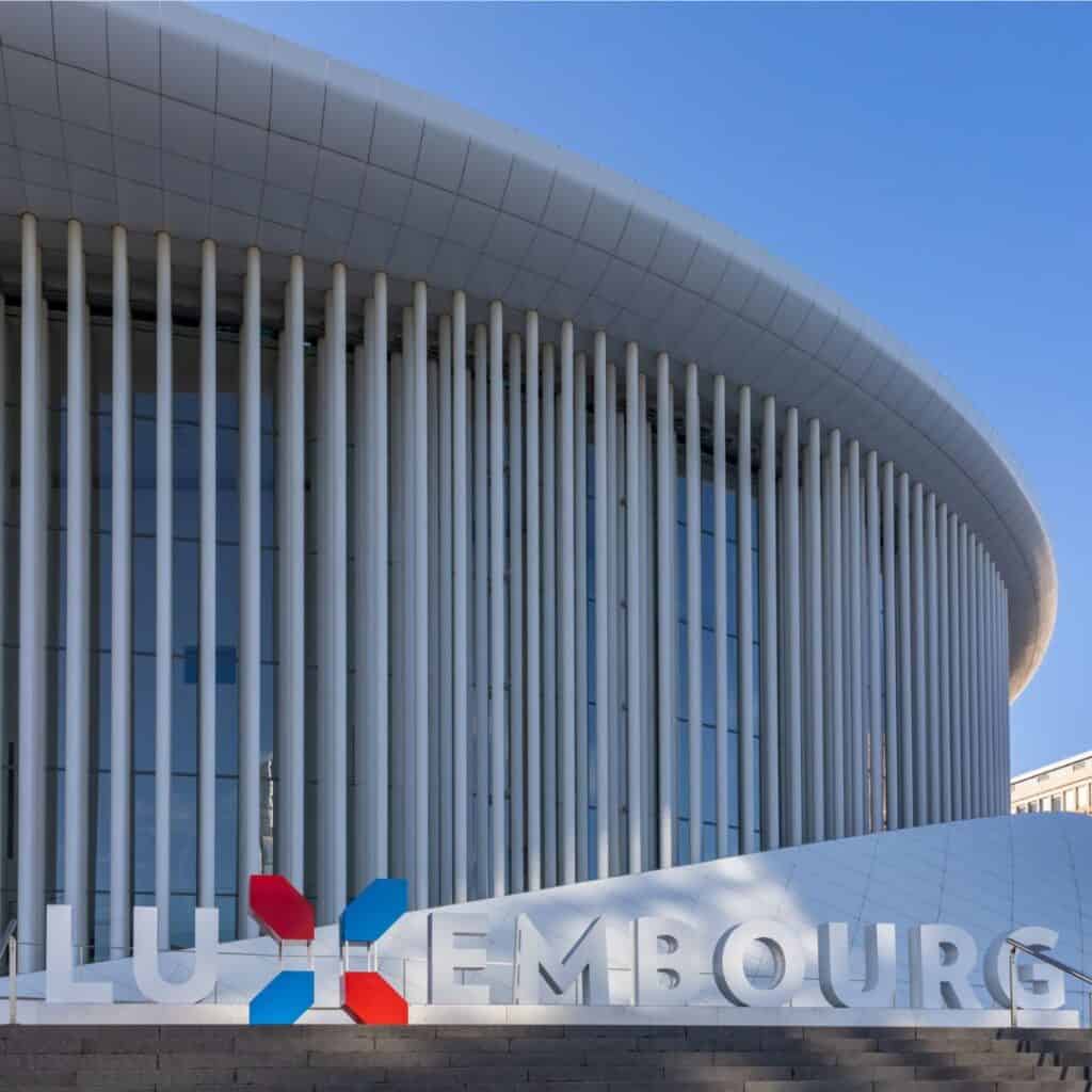 Philharmonie Luxembourg, a modern concert hall building with a glass facade and a curved roof. There is a sign in front of the building that says 