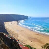 A photo of Praia de Arrifana, a beach in Portugal. The beach is sandy and has a large number of surfers. The waves are crashing against the shore and there are some rocks in the water. The sky is blue and there are some clouds in the distance.