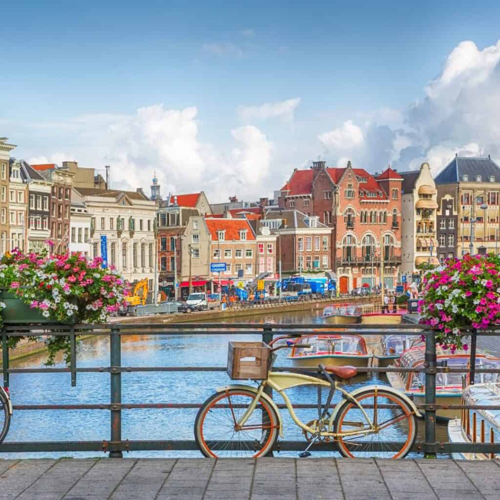 A picturesque view of a canal in Amsterdam, Netherlands. The canal is lined with colorful buildings in various architectural styles, and a flower-filled bridge spans the water. A classic bicycle is parked on the bridge, adding to the charming atmosphere of the scene. The sky is bright blue with some white clouds.