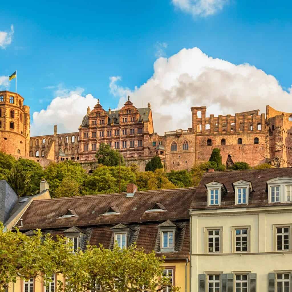 A panoramic view of a Heidelberg Castle, a large, red sandstone structure with multiple towers and buildings, surrounded by lush green trees.