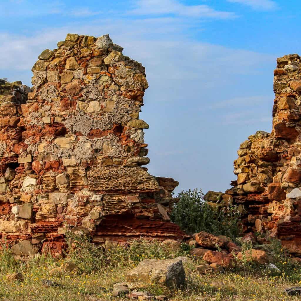 The crumbling ruins of Paderne Castle, with remnants of the walls and foundations still standing. The stones are a mixture of red and brown hues, and there is a lush green field growing around the base of the ruins. The sky is a bright blue, contrasting with the darker tones of the castle.