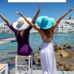 Pinterest pin about Paros Instagram Captions featuring a photo of two women with their arms raised looking out into the view of the ocean in Paros, Greece