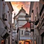 A narrow, cobblestone street in Albufeira, Portugal, lined with whitewashed buildings and adorned with colorful flowers. A church with a prominent cross is visible in the distance, illuminated by the setting sun. The text on the image reads 