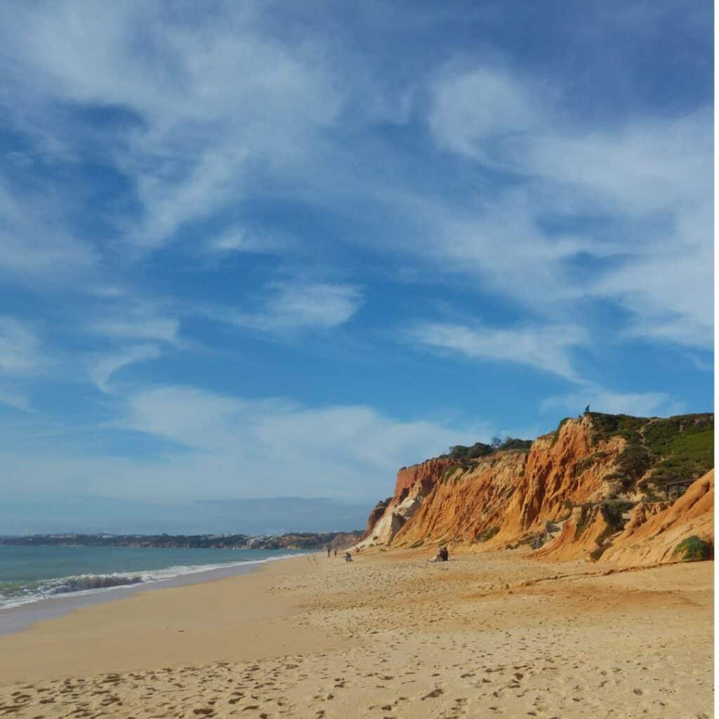 A photo of Praia da Falésia in Algarve, Portugal. The image shows a sandy beach with red cliffs rising in the background. The sky is blue with white clouds. Visiting this place is one of the best things to do in Algarve in September.