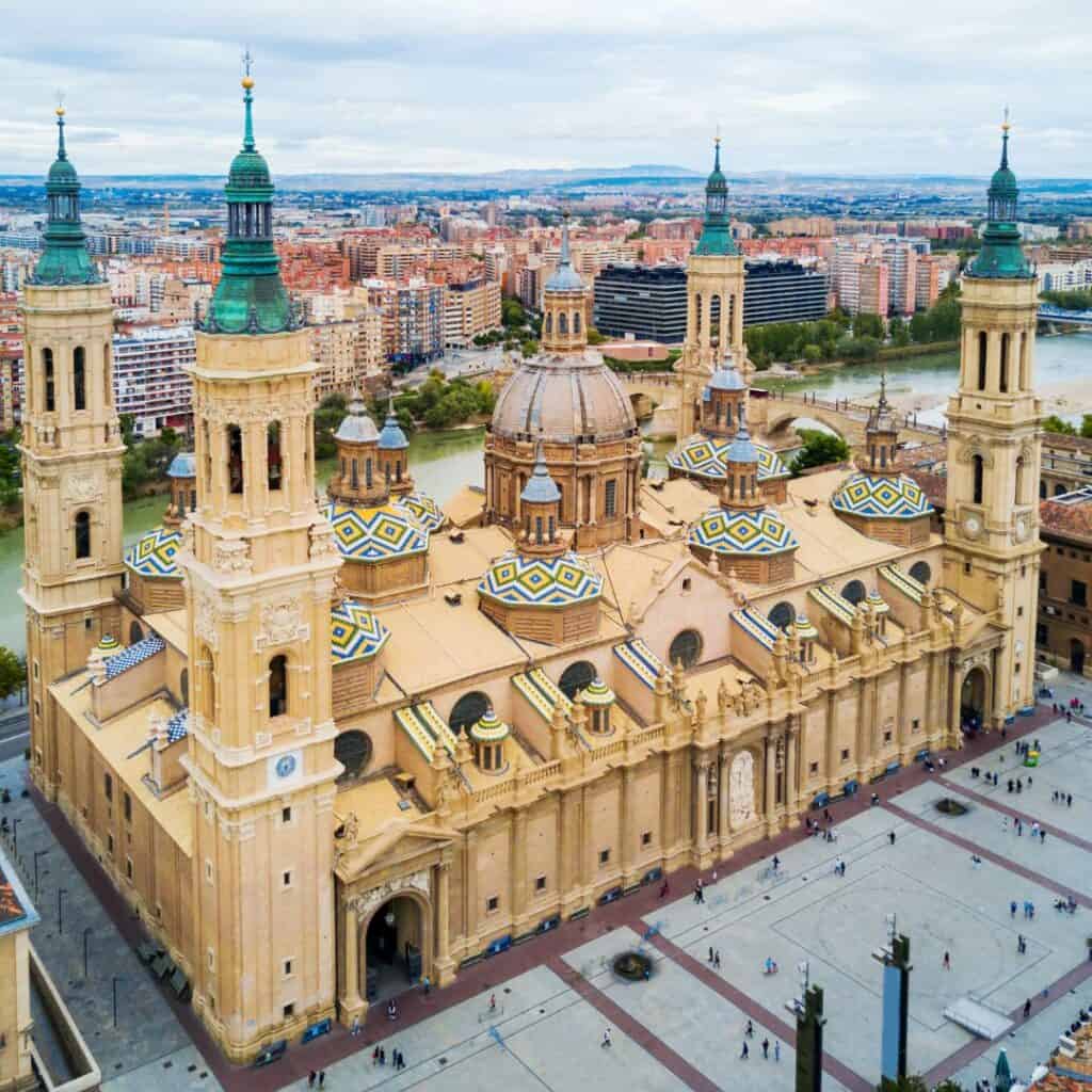 An aerial view of the Basilica of Our Lady of the Pillar in Zaragoza, Spain. The basilica is a large, ornate church with twin towers, domes, and a central courtyard.