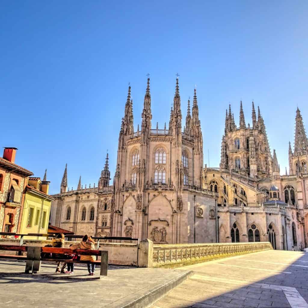 Burgos Cathedral, a large Gothic cathedral in Burgos, Spain, with intricate spires and a bridge leading to its entrance. Two people are sitting on a bench, enjoying the view.