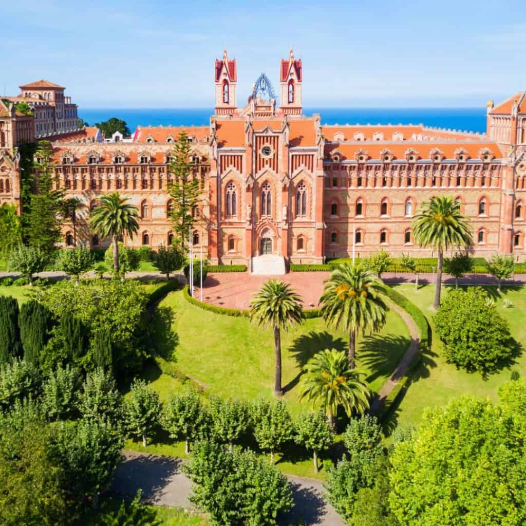 An aerial view of the Pontifical University of Comillas in Comillas, Spain. The university is a large, red-brick building with a central courtyard and palm trees.