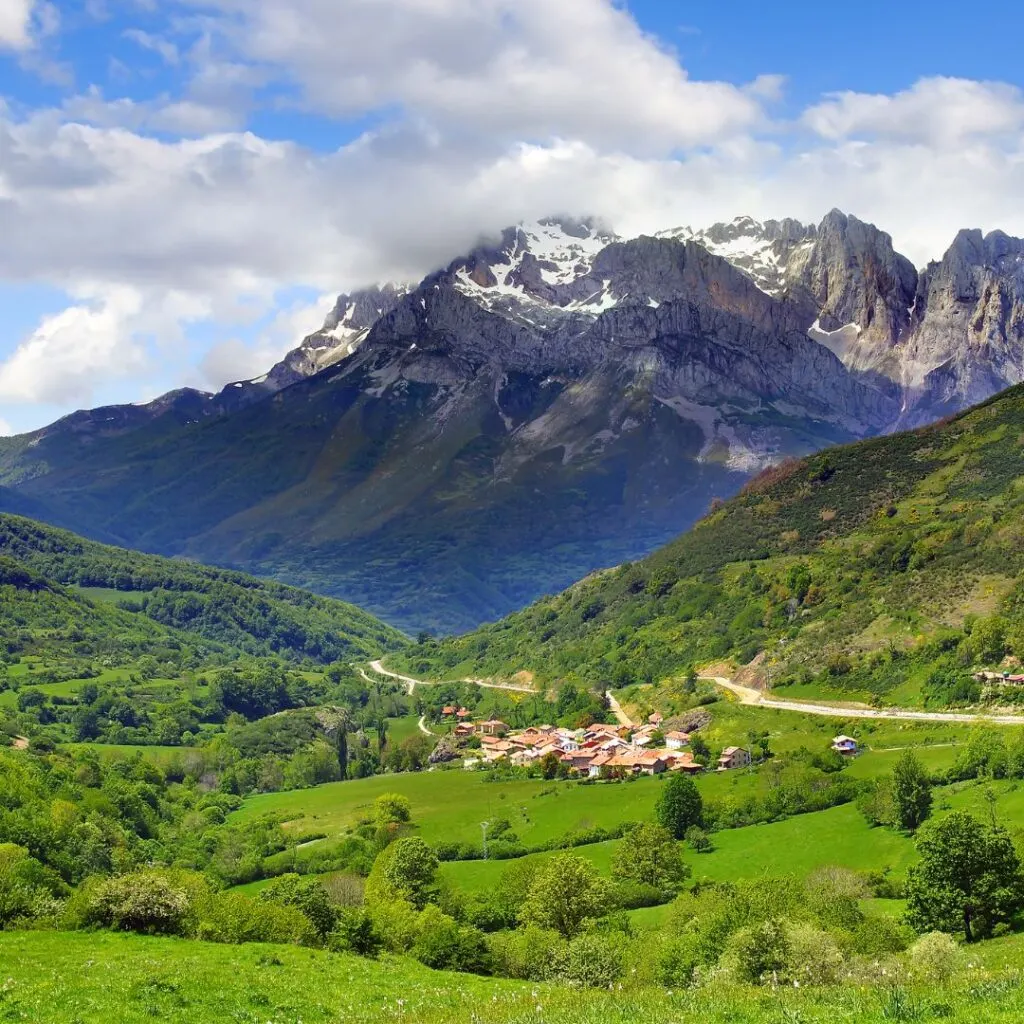 A scenic landscape in the Picos de Europa National Park in Spain, featuring a small village nestled in a valley surrounded by lush green hills and snow-capped mountains.