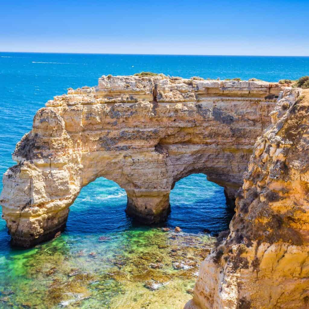  A photo of the Seven Hanging Valleys Trail in Portugal. The image shows a dramatic rock formation with two natural arches overlooking a crystal-clear blue ocean.