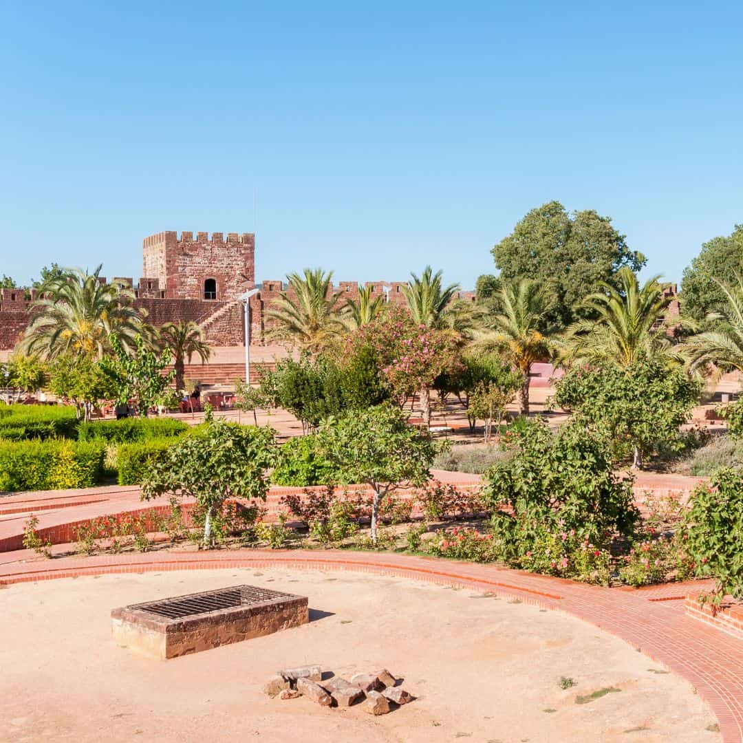 A photo of the Castle of Silves in Algarve, Portugal. The image shows a red-brick castle with a tower, surrounded by palm trees and a landscaped garden.