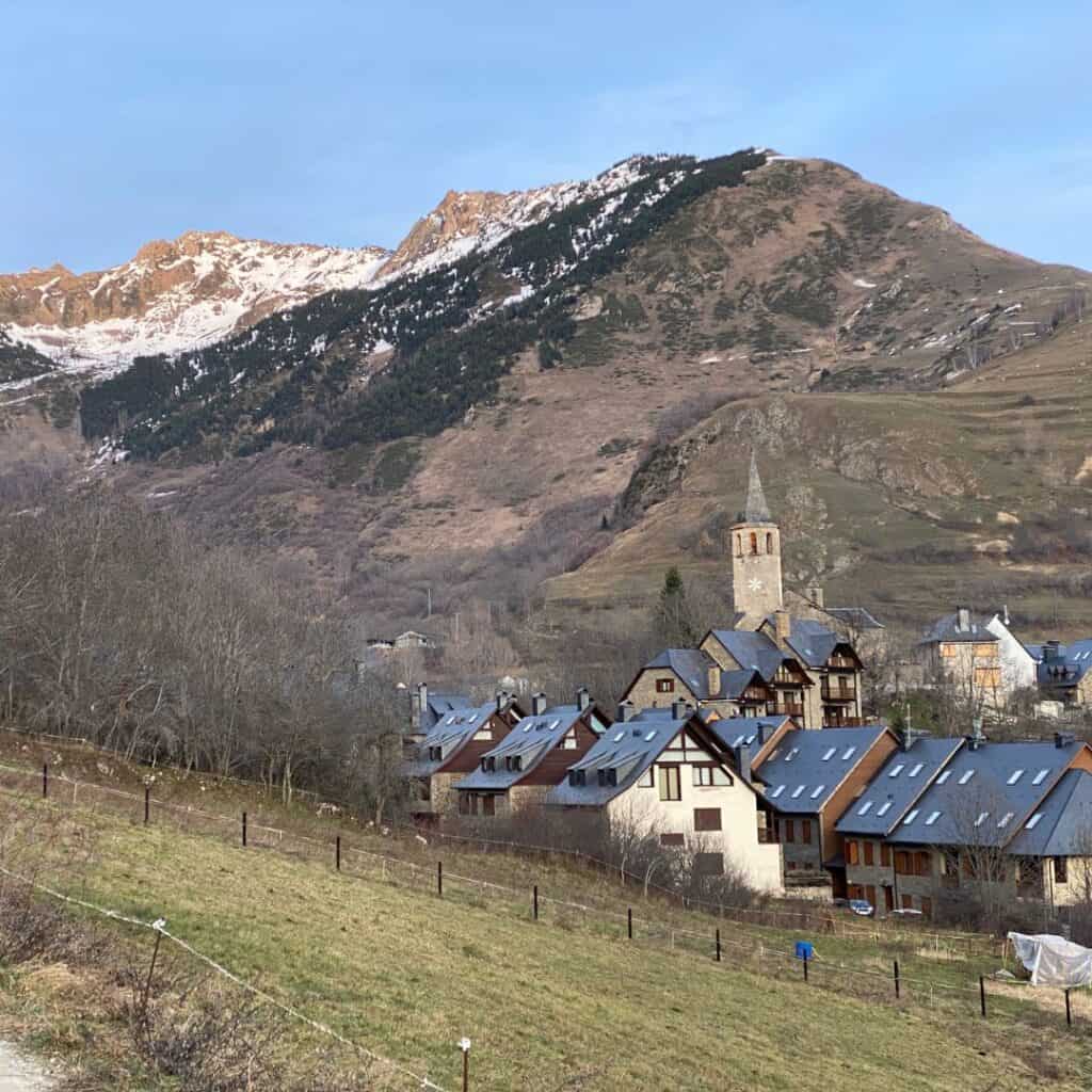 A picturesque village nestled in the Val d'Aran valley in the Pyrenees, with snow-capped mountains in the background and a church tower standing tall above the houses.