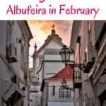 A photo of a narrow cobblestone street lined with whitewashed buildings with red tile roofs in the old town of Albufeira, Portugal. There are a few signs in Portuguese visible on the buildings, including one for a restaurant and a clothing store.