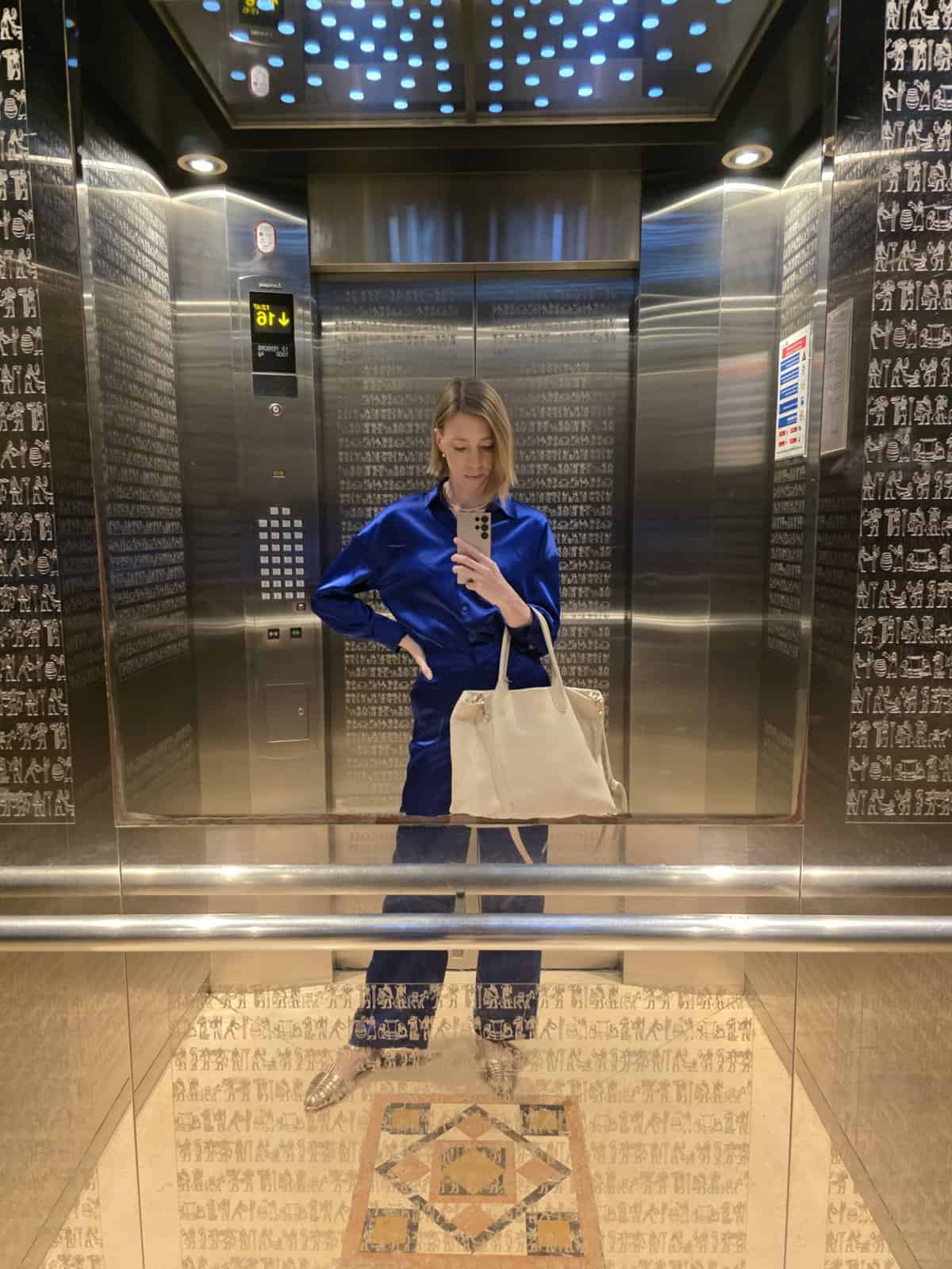 a woman in blue blouse, black pants, and a bag in an elevator taking a picture with a phone
