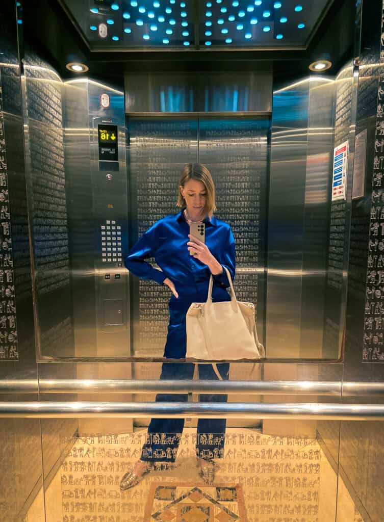 a woman in blue blouse, black pants, and a bag in an elevator taking a picture with a phone