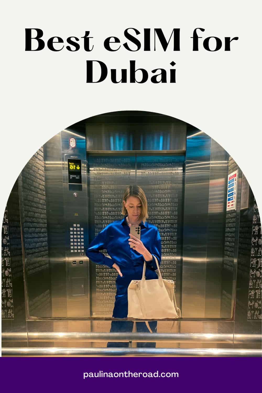 a woman in blue blouse, black pants, and a bag in an elevator taking a picture with a phone